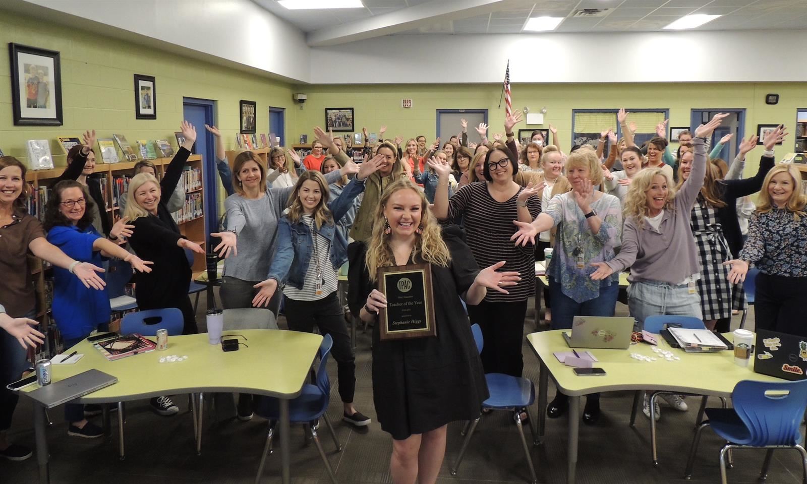 A woman holds a plaque with her coworkers cheering behind her